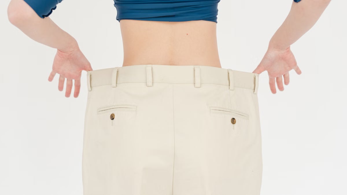 Back view of faceless fit female wearing white oversized trousers standing on white background in light studio after weight loss